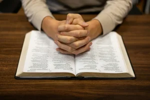 A person sitting at a table with hands folded over a book, symbolizing prayer and reflection on God's grace in difficult times.